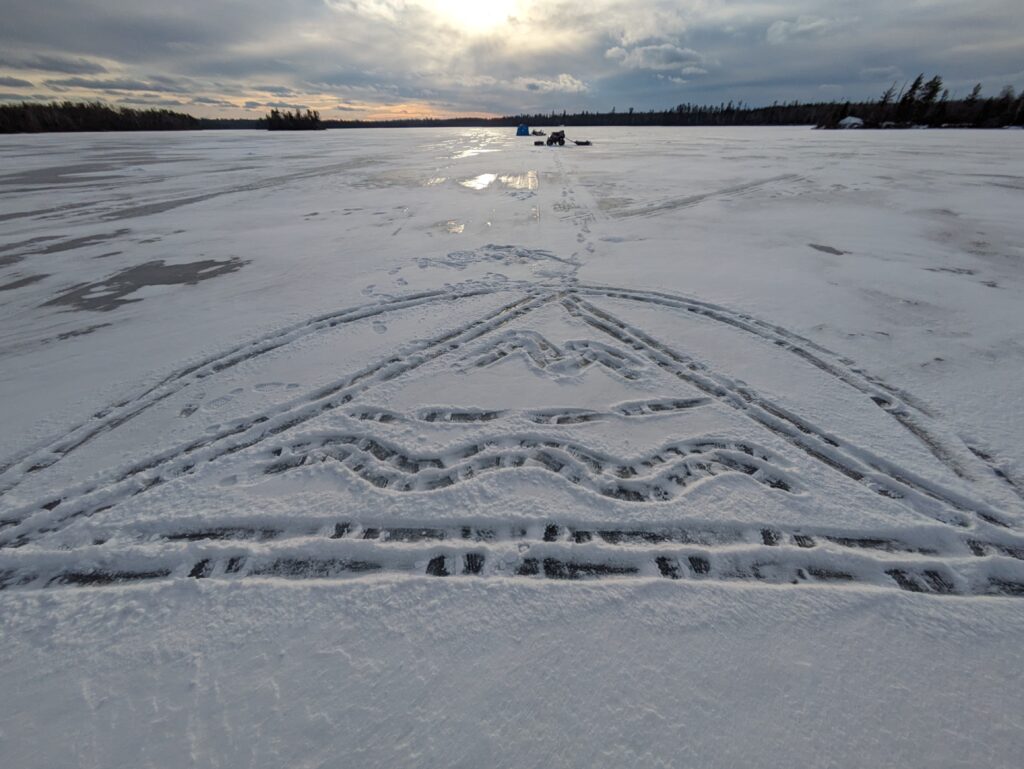 Mosseskogen logo, shuffled into the snow on a frozen lake. The sun is close to setting on the tiny ice fishing crew in the background.